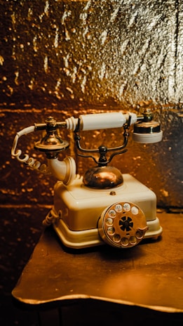 An antique rotary dial telephone with brass and ivory elements rests on a small table. The telephone is designed with intricate metalwork on the cradle and a coiled cord extending from the base. The background features a textured golden surface that adds a rich, warm hue to the scene.
