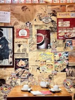 The image features a wall covered with various posters and artwork, including vintage Japanese illustrations and modern advertisements, creating a collage effect. A wooden table in the foreground is set with a teapot, cups, and a menu, suggesting a restaurant or cafe setting. The decor creates a nostalgic and eclectic atmosphere.