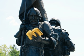 A bronze statue of a medieval knight holding an open gilded book. The knight wears full armor and appears solemn, while a flag is held in the other hand. Another figure, partly visible, stands next to the knight, also in armor.