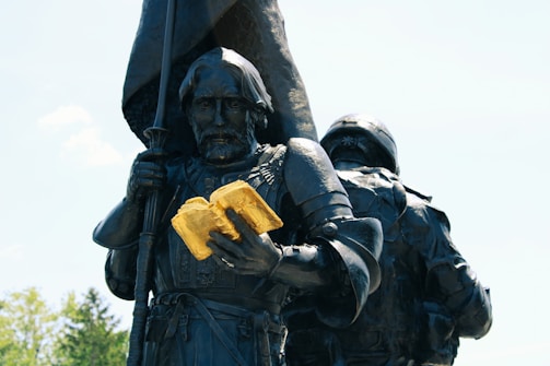 A bronze statue of a medieval knight holding an open gilded book. The knight wears full armor and appears solemn, while a flag is held in the other hand. Another figure, partly visible, stands next to the knight, also in armor.