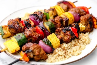 A plate of grilled skewers with chunks of meat, vibrant vegetables such as zucchini, yellow squash, red onion, and red and orange bell peppers. The skewers are arranged over a bed of cooked quinoa, showcasing a combination of charred marks and colorful, fresh ingredients.