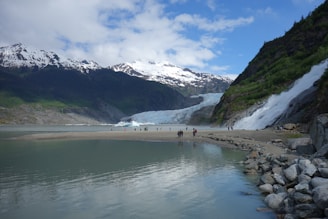 A picturesque landscape with snow-capped mountains in the background, a massive glacier, and a waterfall cascading down into a serene lake. The small group of people at the lake's edge highlights the grand scale of the natural surroundings, including rich green vegetation on the hillsides.