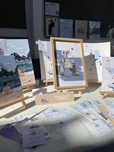 An art display featuring various framed illustrations, including a robot with a heart, beach scenes, and whimsical characters. The display is set up on a table with a bright light casting shadows. A sign identifies the artist as Natasha Guzman, and there are several small cards and prints laid out alongside the frames.