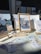 An art display featuring various framed illustrations, including a robot with a heart, beach scenes, and whimsical characters. The display is set up on a table with a bright light casting shadows. A sign identifies the artist as Natasha Guzman, and there are several small cards and prints laid out alongside the frames.