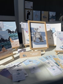 An art display featuring various framed illustrations, including a robot with a heart, beach scenes, and whimsical characters. The display is set up on a table with a bright light casting shadows. A sign identifies the artist as Natasha Guzman, and there are several small cards and prints laid out alongside the frames.
