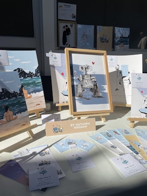 An art display featuring various framed illustrations, including a robot with a heart, beach scenes, and whimsical characters. The display is set up on a table with a bright light casting shadows. A sign identifies the artist as Natasha Guzman, and there are several small cards and prints laid out alongside the frames.
