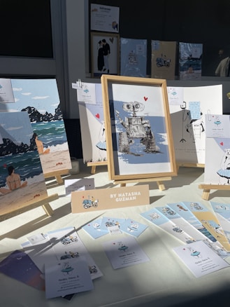 An art display featuring various framed illustrations, including a robot with a heart, beach scenes, and whimsical characters. The display is set up on a table with a bright light casting shadows. A sign identifies the artist as Natasha Guzman, and there are several small cards and prints laid out alongside the frames.