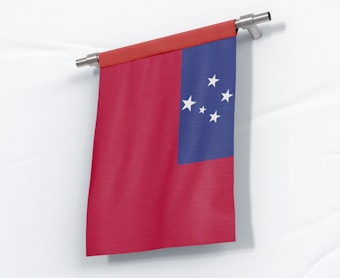 A red wall-mounted flag with a blue canton in the upper left corner containing five white stars arranged in the shape of the Southern Cross constellation. The flag is hanging vertically on a metal rod with brackets, against a white background.