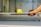 A hand with red nail polish is holding a paddle, balancing a yellow perforated ball. The background includes a blurry indoor sports setting, possibly a tennis or pickleball court.