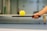 A hand with red nail polish is holding a paddle, balancing a yellow perforated ball. The background includes a blurry indoor sports setting, possibly a tennis or pickleball court.