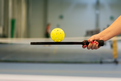 A hand with red nail polish is holding a paddle, balancing a yellow perforated ball. The background includes a blurry indoor sports setting, possibly a tennis or pickleball court.