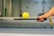 A hand with red nail polish is holding a paddle, balancing a yellow perforated ball. The background includes a blurry indoor sports setting, possibly a tennis or pickleball court.