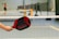 A person holds a black and red pickleball paddle with the brand name 'Conqueror' near a net on a sports court. In the background, there are blurred figures playing on other courts.