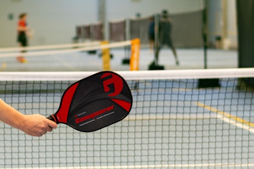 A person holds a black and red pickleball paddle with the brand name 'Conqueror' near a net on a sports court. In the background, there are blurred figures playing on other courts.