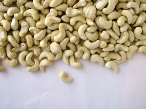 A large pile of shelled cashew nuts spread out on a plain, light-colored surface, with a few scattered away from the main group.