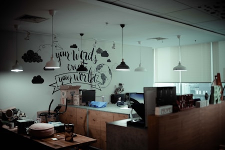 An office space with a mural on the wall that says 'your words create your world.' The room has pendant lights hanging from the ceiling and a window with blinds partially closed. There are multiple desks with computers, various office supplies, and boxes. A wooden partition has decorative items on top, including signs that say 'Home' and 'Hope.'