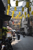A narrow urban street with hanging yellow lanterns overhead. Many motorbikes and scooters are parked and being ridden through the street. A person wearing a traditional conical hat is walking with baskets, possibly selling goods. Signs are visible advertising various services, including currency exchange and SIM cards. Buildings with balconies and foliage overhang the street.