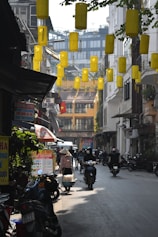 A narrow urban street with hanging yellow lanterns overhead. Many motorbikes and scooters are parked and being ridden through the street. A person wearing a traditional conical hat is walking with baskets, possibly selling goods. Signs are visible advertising various services, including currency exchange and SIM cards. Buildings with balconies and foliage overhang the street.