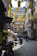A narrow urban street with hanging yellow lanterns overhead. Many motorbikes and scooters are parked and being ridden through the street. A person wearing a traditional conical hat is walking with baskets, possibly selling goods. Signs are visible advertising various services, including currency exchange and SIM cards. Buildings with balconies and foliage overhang the street.