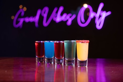 A row of colorful shot glasses filled with different beverages is arranged on a reflective surface. A glowing purple neon sign in the background spells out 'Good Vibes Only.'