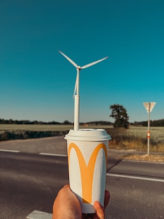 A hand holds a large fast-food beverage cup with a straw, positioned such that a wind turbine in the background visually aligns with the straw. The wind turbine appears to be part of an open landscape with fields and a clear blue sky. A road and some signage are also visible.