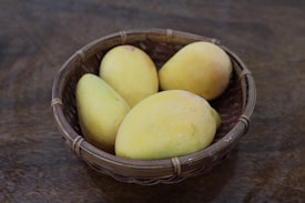 A woven basket containing four ripe yellow mangoes sits on a wooden surface, the natural texture of the wood complementing the smooth skin of the fruit.
