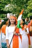 A group of people dressed in white and orange garments are gathered outdoors, holding an orange, white, and green flag. The greenery of trees forms the background, suggesting the image is taken during the day.