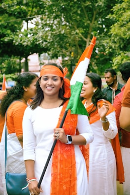 A group of people dressed in white and orange garments are gathered outdoors, holding an orange, white, and green flag. The greenery of trees forms the background, suggesting the image is taken during the day.