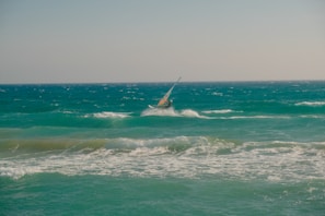 A person is windsurfing on choppy ocean waves under a clear sky. The sea is a deep shade of turquoise with whitecaps visible as the wind pushes against the water. The windsurfer is leaning into the wind, riding the waves actively.