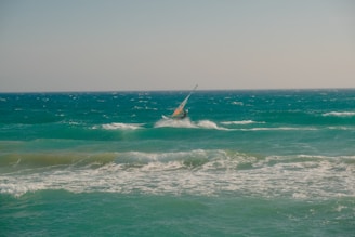 A person is windsurfing on choppy ocean waves under a clear sky. The sea is a deep shade of turquoise with whitecaps visible as the wind pushes against the water. The windsurfer is leaning into the wind, riding the waves actively.