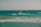 A person is windsurfing on choppy ocean waves under a clear sky. The sea is a deep shade of turquoise with whitecaps visible as the wind pushes against the water. The windsurfer is leaning into the wind, riding the waves actively.