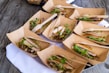 Several cardboard containers are arranged on a wooden surface, each containing a serving of food that includes slices of meat, microgreens, and grains. Wooden forks are placed in some of the containers. The containers are positioned on a white cloth.