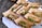 Several cardboard containers are arranged on a wooden surface, each containing a serving of food that includes slices of meat, microgreens, and grains. Wooden forks are placed in some of the containers. The containers are positioned on a white cloth.