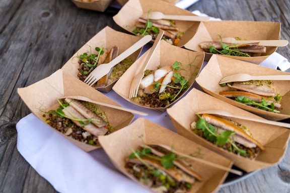 Several cardboard containers are arranged on a wooden surface, each containing a serving of food that includes slices of meat, microgreens, and grains. Wooden forks are placed in some of the containers. The containers are positioned on a white cloth.