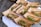 Several cardboard containers are arranged on a wooden surface, each containing a serving of food that includes slices of meat, microgreens, and grains. Wooden forks are placed in some of the containers. The containers are positioned on a white cloth.