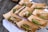 Several cardboard containers are arranged on a wooden surface, each containing a serving of food that includes slices of meat, microgreens, and grains. Wooden forks are placed in some of the containers. The containers are positioned on a white cloth.