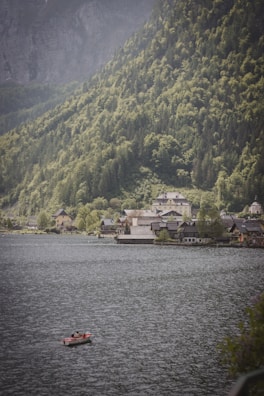 A picturesque lakeside town nestled against lush, green mountains. In the foreground, a small red boat with a person inside drifts on the lake's calm, rippling surface. The background is dominated by steep, verdant hills and scattered houses with traditional architecture along the shore.