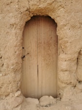 A rustic, wooden door is set within a textured, earthy wall made of a clay-like material. The door is simple and weathered, with visible grains running vertically. The surrounding wall has an uneven and coarse surface, giving an ancient and sturdy appearance.