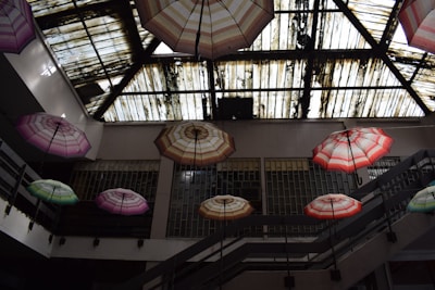 Colorful umbrellas are suspended from the ceiling inside a building with a partially transparent roof. The roof appears to be old and weathered, showing streaks of dirt and grime. The umbrellas are in varying shades of pink, green, and red, creating a vibrant contrast against the muted tones of the interior walls and railings.