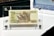 An Indonesian banknote of 5000 Rupiah displayed in a transparent protective case. The note features intricate designs and text, including 'BANK INDONESIA' and 'LIMA RIBU RUPIAH'. There is a detailed illustration of traditional Indonesian architecture on the note.