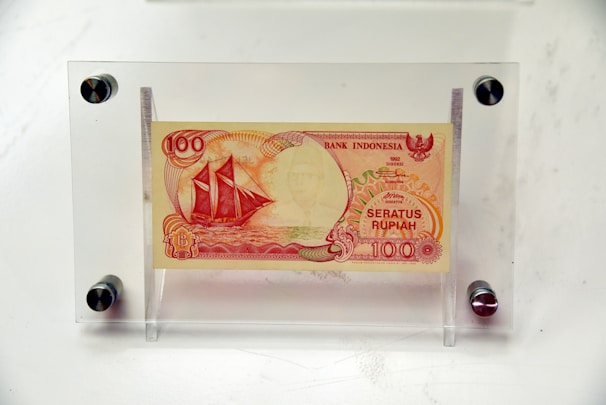 A 100 Rupiah Bank Indonesia note from 1992 is enclosed in a clear protective display case, with a design featuring an image of a traditional sailing ship and intricate patterns in red and orange hues.