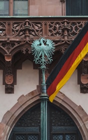 An ornate bronze sculpture of a double-headed eagle is mounted on top of a tall pole, set against the backdrop of an intricate stone facade. A German flag is prominently displayed, with its black, red, and yellow colors.