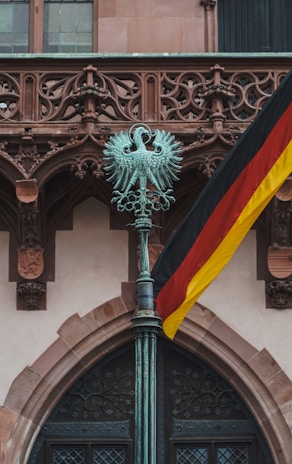 An ornate bronze sculpture of a double-headed eagle is mounted on top of a tall pole, set against the backdrop of an intricate stone facade. A German flag is prominently displayed, with its black, red, and yellow colors.