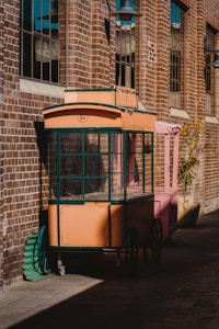 A vintage-style cart with an orange body and green trim is positioned against a brick wall, partially shaded. The cart is on wheels and appears to be an old market or vendor cart. There are large windows on the building, casting shadows on the ground. A pink structure is adjacent, and a round green plate leans against the cart.