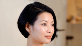 A portrait of a person with a short, sleek black hairstyle, wearing a subtle makeup with glossy lips and a serene expression. The background is softly blurred and neutral in color.