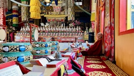 An ornate and vividly decorated interior of a monastery or temple is depicted, with intricate patterns, vibrant colors, and religious artifacts. A monk sits on a raised platform to the right, dressed in traditional robes and appears engaged in prayer or study, surrounded by religious texts, instruments, and ceremonial objects. The scene is filled with decorative elements including fabric hangings, religious icons, and detailed murals.