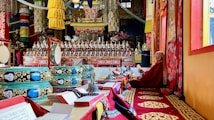 An ornate and vividly decorated interior of a monastery or temple is depicted, with intricate patterns, vibrant colors, and religious artifacts. A monk sits on a raised platform to the right, dressed in traditional robes and appears engaged in prayer or study, surrounded by religious texts, instruments, and ceremonial objects. The scene is filled with decorative elements including fabric hangings, religious icons, and detailed murals.