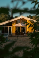 A yellow wooden house with a slanted roof is surrounded by trees and greenery. The house has large windows and a patio area with chairs and a parasol. The scene is framed by blurred leaves in the foreground, suggesting a secluded, peaceful setting.