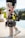A person holding a professional video camera with a gimbal stabilizer, wearing light-colored pants and white sneakers. The person appears to be walking outdoors on a sunny day, with a blurred background of foliage and distant landscape.