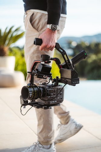 A person holding a professional video camera with a gimbal stabilizer, wearing light-colored pants and white sneakers. The person appears to be walking outdoors on a sunny day, with a blurred background of foliage and distant landscape.